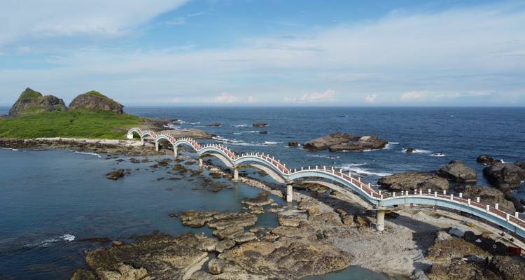 Puente peatonal curvado de múltiples arcos que conecta islotes rocosos sobre un mar azul profundo.