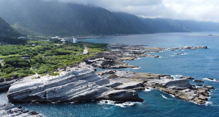 Vista aérea de una costa escarpada con laderas boscosas que se encuentran con aguas turquesas bajo nubes dispersas.