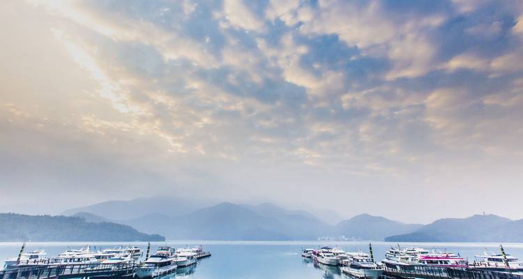 Marina de barcos turísticos amarrados en un lago plácido bajo un cielo dramático nublado y montañas brumosas.