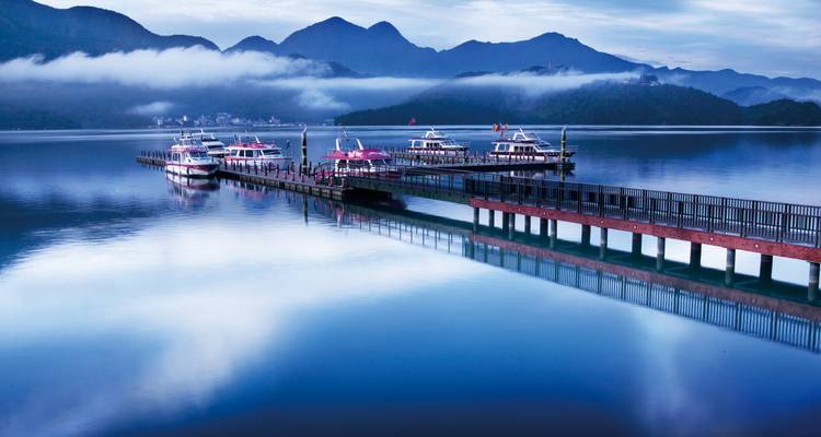 Muelle de madera extendiéndose hacia un lago azul cristalino con botes amarrados y nubes bajas abrazando las crestas de las montañas.
