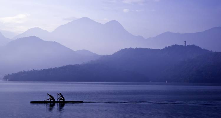 Remeros en silueta en una canoa estrecha surcando un lago brumoso ante picos montañosos escalonados.