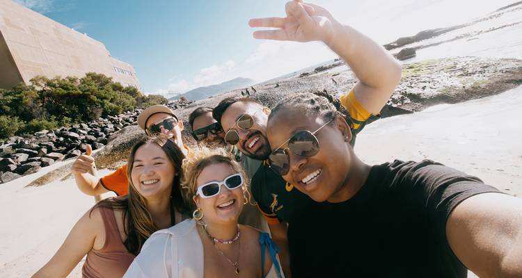 Een vrolijke groep maakt een selfie op een zonnig strand met rotsachtige kustlijn en verre bergen.