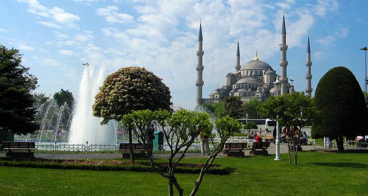 View across a fountain-filled park toward the silhouette of the Blue Mosque under a lightly clouded sky.
