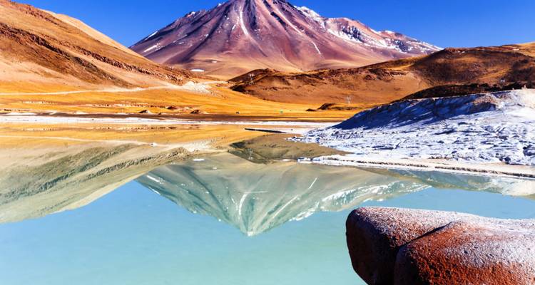 Una laguna espejada refleja un imponente volcán andino bajo un cielo azul profundo.