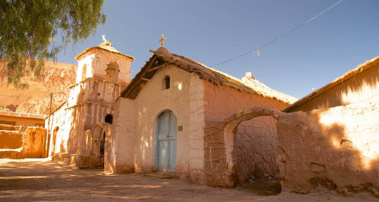 Iglesia de adobe rústica con campanario y arco situados contra un telón de fondo de montañas áridas.
