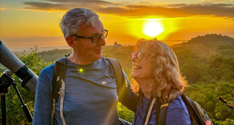 Una pareja sonriente comparte un momento de amanecer en un mirador de la selva con la silueta de un templo antiguo a lo lejos.