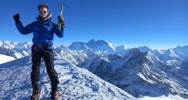Persoon die een ijsbijl vasthoudt op een besneeuwde bergtop met een uitgestrekt berglandschap op de achtergrond.