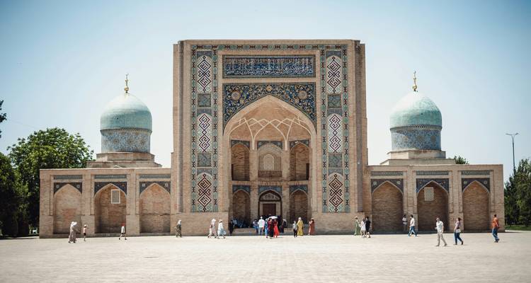 A mosque with stone domes, a large arch, and visitors in the foreground.