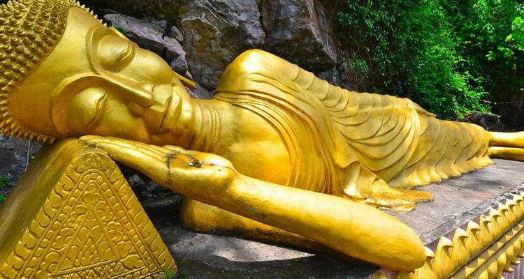 Goldene liegende Buddha-Statue vor einem Hintergrund aus Felsen und grünem Laub.