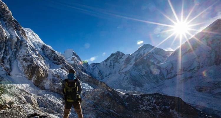Person, die auf einem felsigen Berg mit schneebedeckten Gipfeln und einer strahlenden Sonne am Himmel steht.