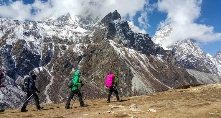 Drei Wanderer mit Trekkingstöcken gehen entlang eines Bergpfades mit schneebedeckten Gipfeln im Hintergrund.