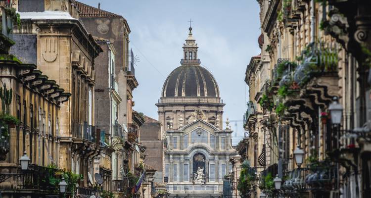 La cúpula de la catedral barroca domina una calle estrecha llena de balcones ornamentados y plantas en Sicilia.