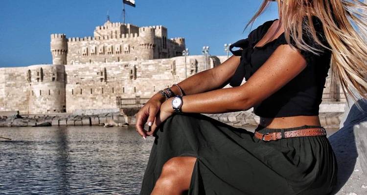 Jeune femme posant près de la mer avec les murs en pierre du Fort Qaitbay derrière elle.