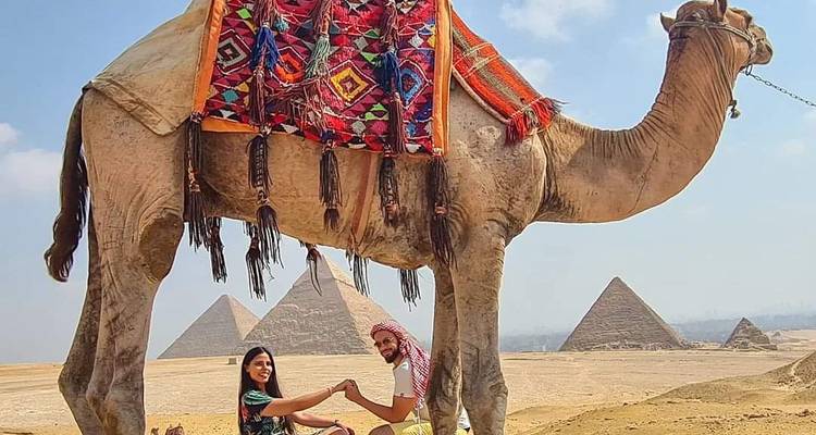 Un chameau au premier plan encadre un couple se tenant par la main avec les pyramides derrière eux.