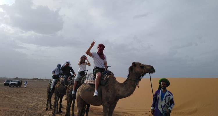 Menschen reiten auf Kamelen durch eine Wüstenlandschaft unter einem bewölkten Himmel.