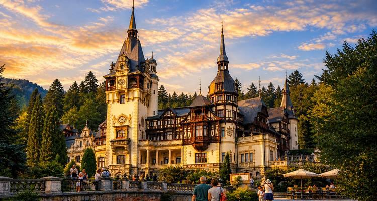 Peles Castle bathed in golden sunset light, nestled among dense forested hills with tourists admiring the view.