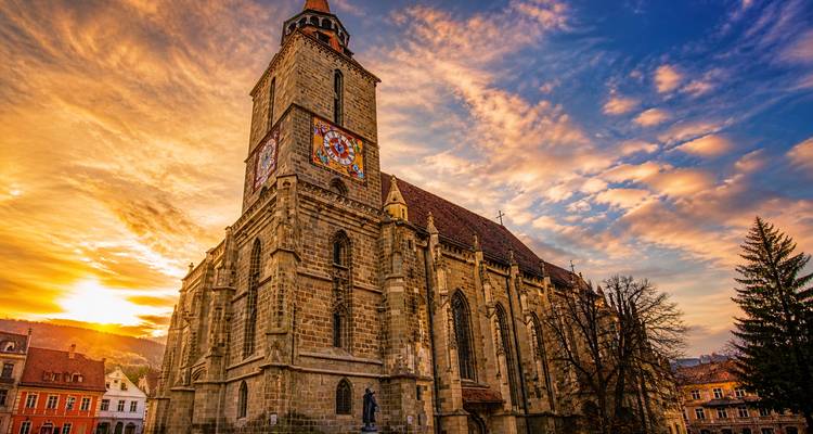 The Black Church of Brasov rises against a dramatic orange-pink sunset sky, its gothic tower and stained-glass clock vividly illuminated.
