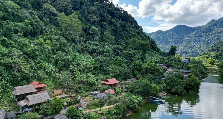 Perspectiva de dron de pueblos ribereños sobre pilotes y laderas cubiertas de selva que se encuentran con aguas verdes tranquilas.