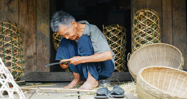 Artesano anciano se agacha mientras talla tiras de bambú para tejer cestas tradicionales afuera de una casa de madera.
