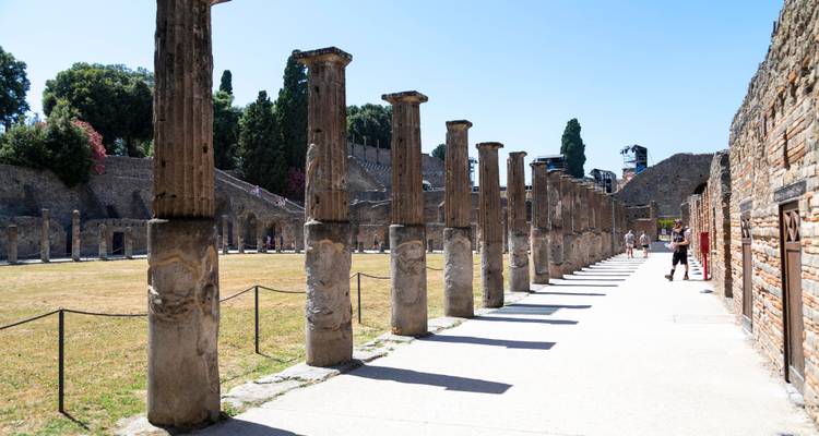 Fila de columnas en pie proyectando sombras en la palestra de Pompeya con visitantes caminando por el sendero.