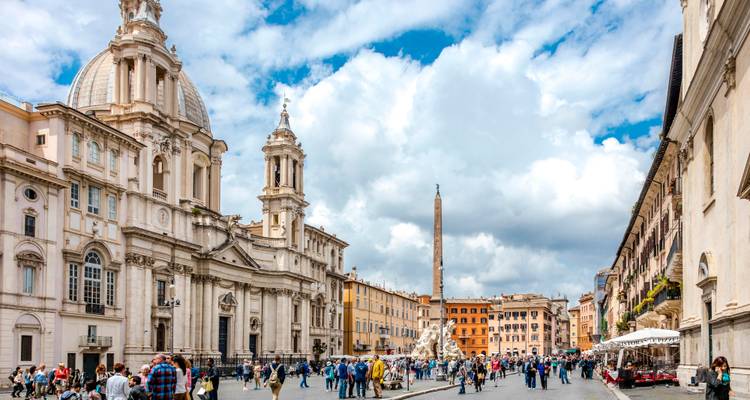 La bulliciosa Piazza Navona rodeada de arquitectura barroca, cafés y la fuente con obelisco central en un día soleado.