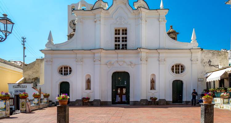 Elegante fachada de iglesia de estilo barroco blanco dando hacia una plaza de ladrillo abierta con flores en macetas bajo un cielo azul despejado.
