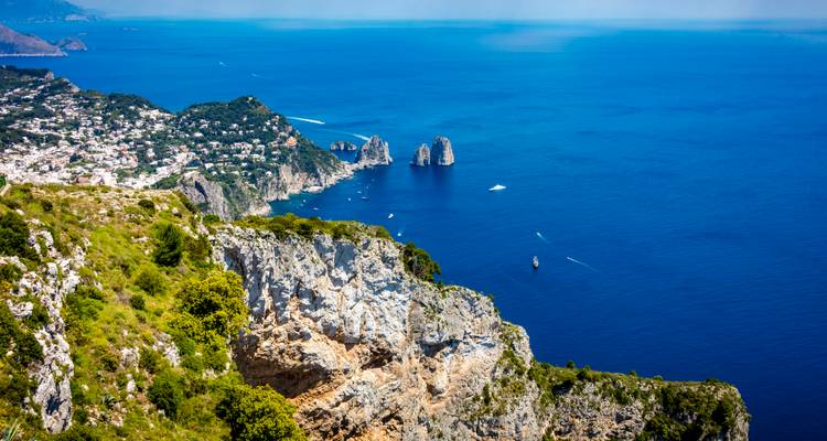 Acantilado dramático que cae hacia el mar azul profundo con las rocas Faraglioni y el pueblo de Capri abajo.