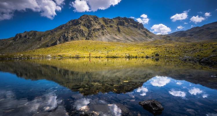 Un lac alpin élevé reflétant parfaitement une crête montagneuse escarpée avec un randonneur solitaire au loin.