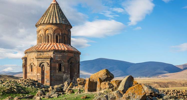 Une église en pierre isolée à tour ronde se dresse sur une plaine rocheuse avec des montagnes au loin sous un ciel partiellement nuageux.