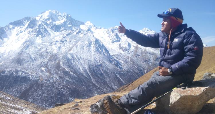 Eine Person, die auf einem Felsen sitzt und einen Wanderstock umklammert, vor einer Bergkulisse mit schneebedeckten Gipfeln.