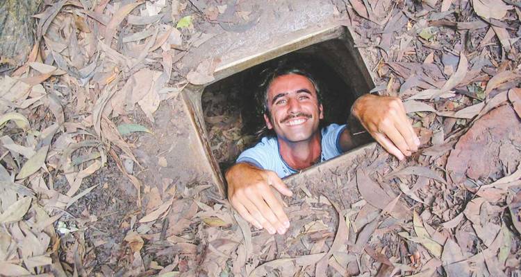 Un voyageur souriant sort d'une petite entrée de tunnel carrée située dans le sol de la forêt, avec des feuilles sèches tout autour.