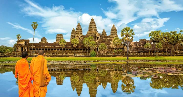 Two monks in orange robes walking by Angkor Wat with a reflection in the water.