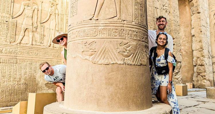 Groupe de touristes souriants qui regardent derrière une colonne sculptée d'hiéroglyphes à l'intérieur d'un temple égyptien antique.
