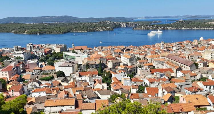 Vogelperspektive über die Terrakotta-Dächer von Sibenik in Richtung des tiefblauen Kanals mit einem Kreuzfahrtschiff