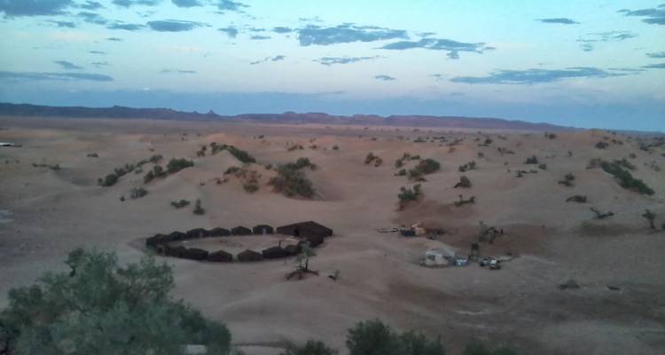 Une vue large d'un campement circulaire du désert composé de tentes sombres installées parmi des dunes de sable ondulantes sous un ciel du soir bleu qui s'estompe.
