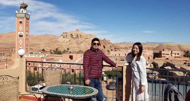 Deux voyageurs posent sur une terrasse de toit surplombant une ville du désert avec un grand minaret et des collines lointaines sous un ciel bleu clair.