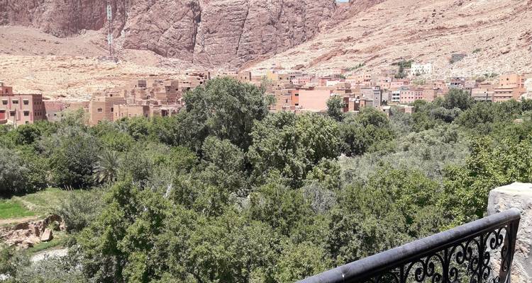 Une oasis luxuriante d'arbres verts borde un village couleur ocre adossé à de hautes falaises rocheuses sous une lumière vive de milieu de journée.