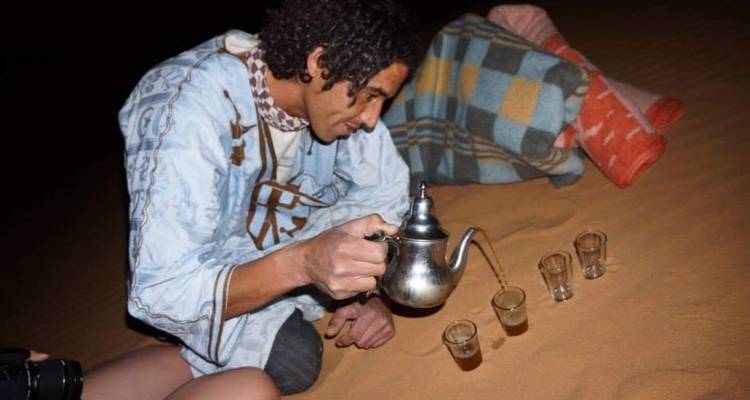 Un jeune homme en robe traditionnelle verse du thé à la menthe dans de petits verres alors qu'il est assis sur le sable du désert la nuit.