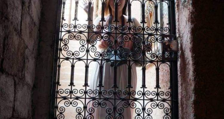 Une femme en robe claire se tient derrière un portail en fer forgé orné avec la lumière du soleil qui filtre à travers.