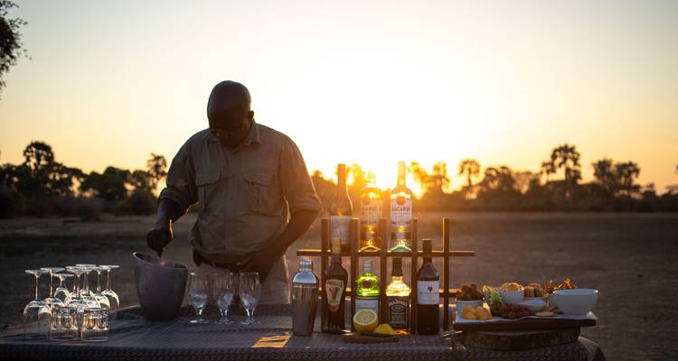 Un guide prépare des apéritifs au coucher du soleil à une table de bar en brousse se détachant en silhouette contre un coucher de soleil orangé