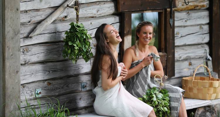 Two people laughing and relaxing in towels outside a wooden sauna.