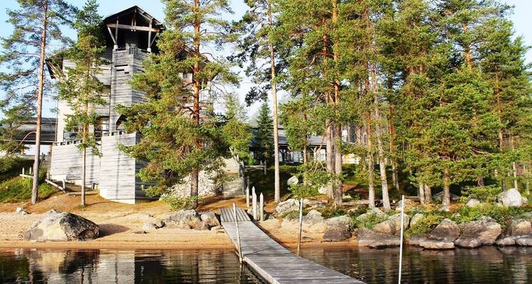 A wooden tower and lodge surrounded by pine trees by a lake.