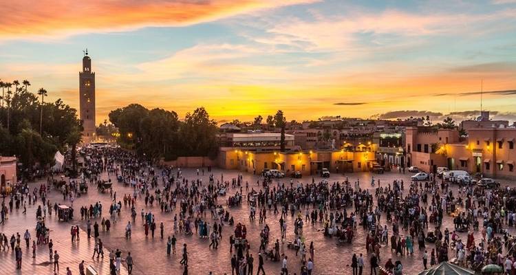 Een druk stadsplein bij zonsondergang, met talrijke mensen en een hoge toren op de achtergrond.