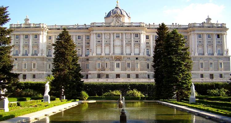Het elegante Koninklijk Paleis van Madrid omgeven door cipressen en een spiegelend waterbekken op een zonnige middag.