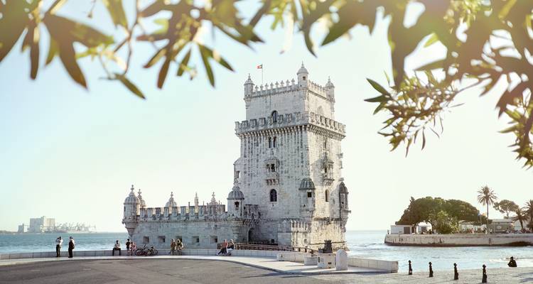 La Torre de Belém se dresse aux côtés du Tejo encadrée par des branches feuillues avec quelques visiteurs sur la promenade.