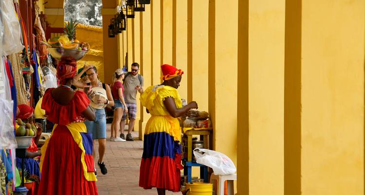 Arcade jaune vibrant à Carthagène où des femmes Palenqueras vêtues de couleurs vives vendent des fruits au passage des touristes