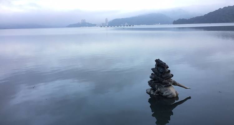 Eaux matinales calmes du lac Sun Moon avec un horizon brumeux et un petit cairn de pierre au premier plan.