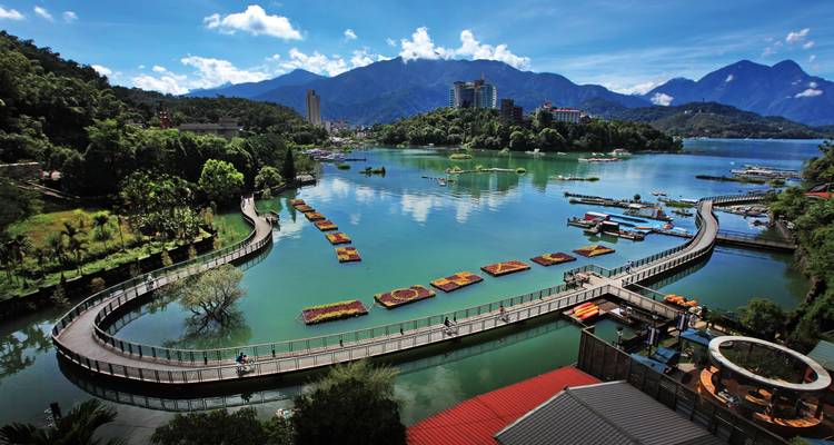Vue aérienne d'une promenade de bord de lac sinueuse et de parterres de fleurs flottants avec horizon urbain et montagnes environnantes.