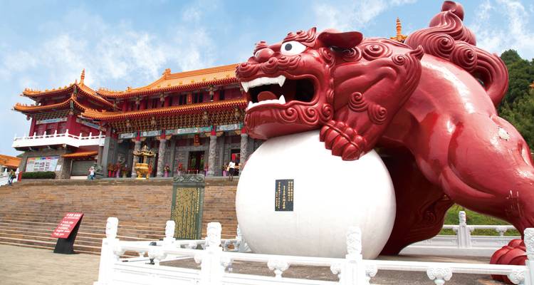 Groot rood standbeeld van een mythisch wezen buiten een traditioneel Chinees gebouw.