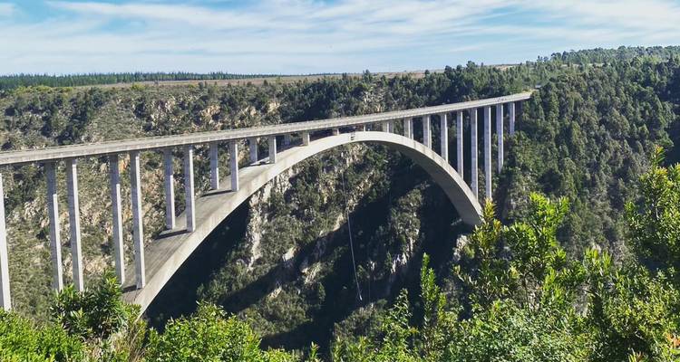 Un grand pont en arc de béton enjambe une gorge boisée profonde sous un ciel bleu vif avec des nuages effilés.